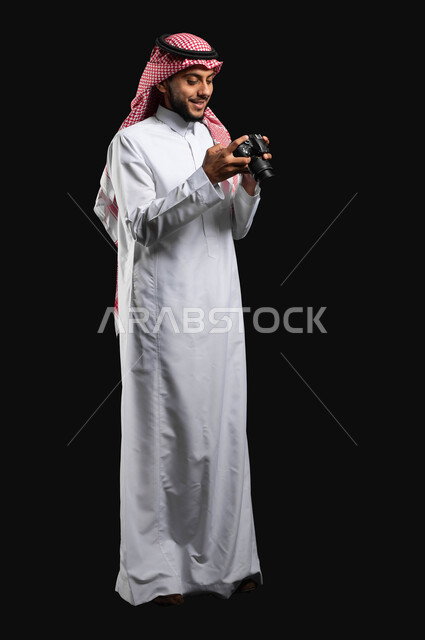مفهوم التصوير الفوتوغرافي, بورتريه لشاب عربي خليجي سعودي يرتدي الزي التقليدي والشماغ، يحمل بيده كاميرا التصوير وينظر إلى الصور الملتقطة  ويبتسم، موهبة واحتراف فن التصوير ، مصورين سعوديين ، كاميرات عالية الدقة, مجال التصوير والإعلام, الجسم بالطول الكامل