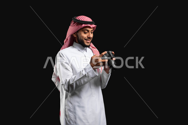 استخدام التكنولوجيا في مجال التصوير , بورتريه بخلفية سوداء  لشاب عربي خليجي سعودي يرتدي الزي التقليدي والشماغ، يحمل بيده كاميرا التصوير وينظر إلى الصور الملتقطة ، التصوير الفوتوغرافي ، موهبة واحتراف فن التصوير ، مصورين  محترفيين سعوديين