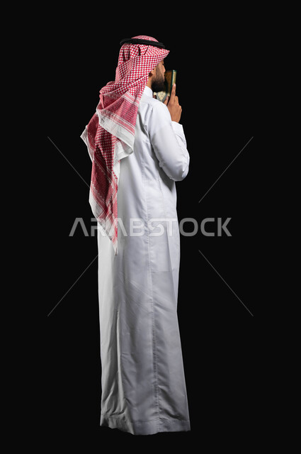 Performing the duties of Islam, a portrait with a black background of a Saudi Gulf Arab man wearing the traditional white thobe and the Saudi shemagh, holding the Holy Qur’an in his hands and kissing it, remembrance of God Almighty, seeking forgiveness and supplication, humility and supplication to God, full-length body, Islamic worship and rituals