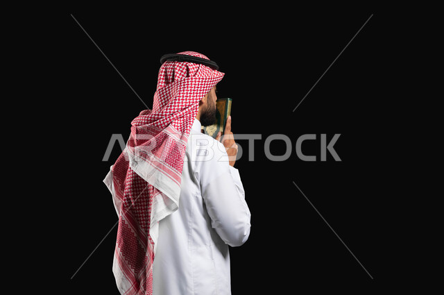 Humility in performing acts of worship, a portrait with a black background of a Saudi Gulf Arab man wearing the traditional white thobe and the Saudi shemagh, holding the Holy Qur’an in his hands and kissing it, remembrance of God Almighty, glorifying the sanctities of Islam, seeking forgiveness and supplication, diligence in worship