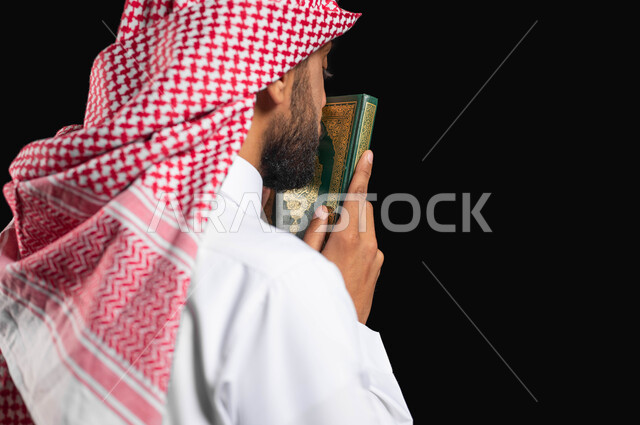 تعظيم المقدسات الإسلامية, بورتريه مقرب من الجانب لرجل عربي سعودي خليجي يرتدي الثوب الأبيض التقليدي والشماغ السعودي, يمسك القرآن الكريم بيديه ويقبله, الإكثار من ذكر الله تعالى, الإستغفار والدعاء, الإجتهاد في العبادة, خلفية سوداء