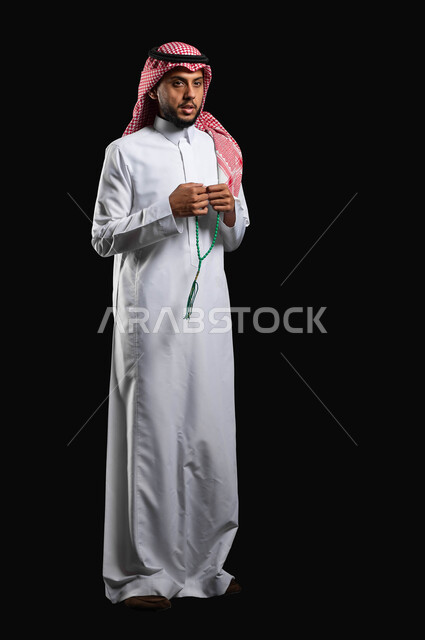 التسبيح والتهليل وذكر الله تعالى، الاستغفار والصلاة على النبي, الإجتهاد في العباده، بورتريه لرجل عربي سعودي خليجي يرتدي الثوب والشماغ التقليدي يمسك بيده السبحه, قراءة أذكار ما بعد الصلاة، الجسم بالطول الكامل، خلفية سوداء، استخدام حساس، محتوى حساس