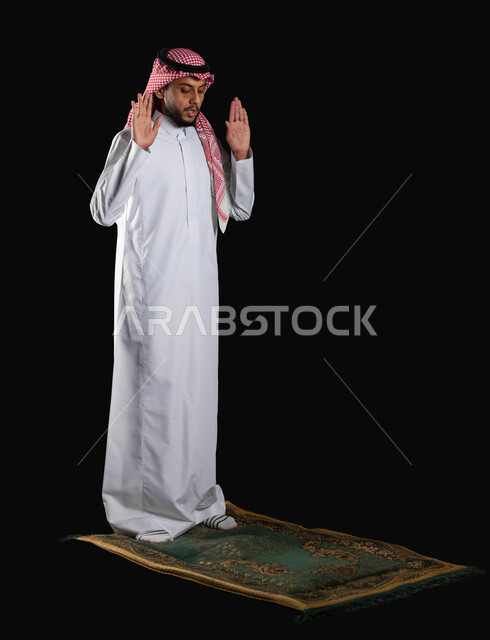 الالتزام بأداء الفروض على وقتها، ايماءات التكبير لبدء الصلاة، الركن الثاني من اركان الاسلام، بورتريه لرجل عربي خليجي سعودي مسلم يرتدي الثوب التقليدي والشماغ يقف على السجادة، العبادة والتقرب إلى الله، الجسم بالطول الكامل، خلفية سوداء، استخدام حساس، محتوى حساس