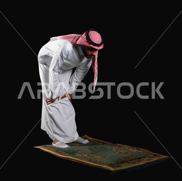الاجتهاد في العباده والطاعة، الركن الثاني من اركان الاسلام، التقرب إلى الله عن طريق القيام بالفرائض والعبادات، بورتريه لرجل عربي خليجي سعودي يرتدي الثوب التقليدي والشماغ يقوم بالركوع أثناء الصلاة، الجسم بالطول الكامل، خلفية سوداء, استخدام حساس, محتوى حساس