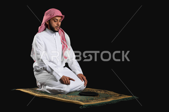 العبادة و التقرب الى الله، رفع اصبع السبابة للأعلى للتشهد، أهمية الالتزام بالصلاة على وقتها، بورتريه من الجانب لرجل عربي سعودي خليجي يرتدي الثوب التقليدي والشماغ يجلس على سجادة الصلاة، خلفية سوداء، محتوى حساس، استخدام حساس