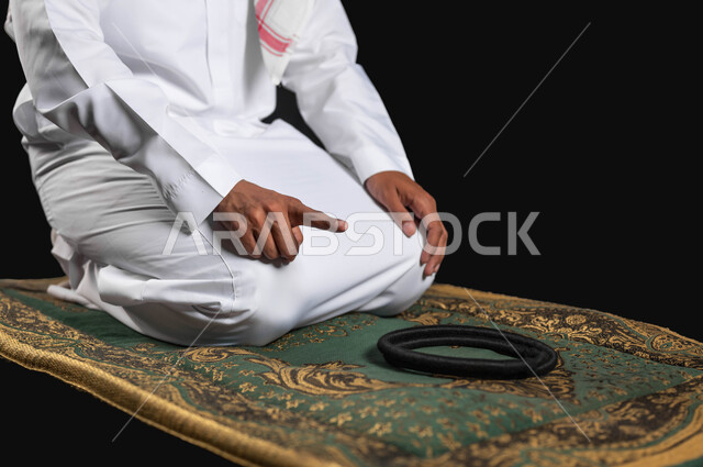 أهمية الالتزام بالصلاة على وقتها، بورتريه مقرب ليد رجل عربي سعودي خليجي يرتدي الثوب التقليدي والشماغ يجلس على سجادة الصلاة، العبادة و التقرب الى الله يرفع اصبع السبابة للأعلى للتشهد، خلفية سوداء، محتوى حساس، استخدام حساس