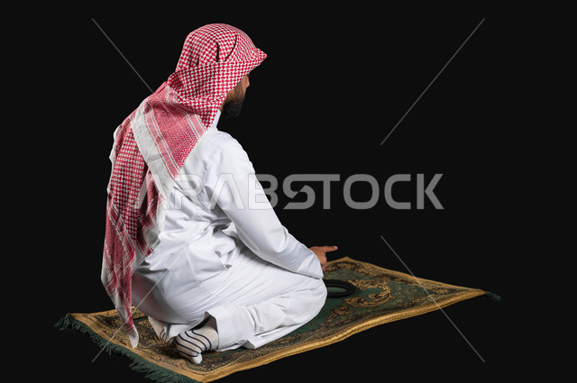 رفع اصبع السبابة للأعلى للتشهد، أهمية الالتزام بالصلاة على وقتها، بورتريه من الجانب لرجل عربي سعودي خليجي يرتدي الثوب التقليدي والشماغ يجلس على سجادة الصلاة، العبادة و التقرب الى الله، خلفية سوداء، محتوى حساس، استخدام حساس