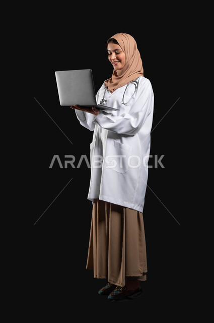 مفهوم استخدام أجهزة تقنيه حديثه، العمل في القطاع الصحي، بورتريه لطبيبة عربية خليجية سعودية مبتسمه محجبة ترتدي العباءه مع المعطف الطبي تحمل بيدها الحاسوب المحمول, صورة الجسم بالطول الكامل، خلفيه سوداء، محتوى حساس، استخدام حساس
