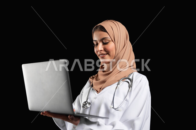 العمل في القطاع الصحي، مفهوم استخدام أجهزة تقنيه حديثه، بورتريه مقرب لطبيبة عربية خليجية سعودية مبتسمه محجبة ترتدي المعطف الطبي تحمل بيدها الحاسوب المحمول, خلفيه سوداء، محتوى حساس، استخدام حساس