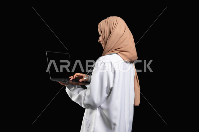 مفهوم استخدام أجهزة تقنيه حديثه، العمل في القطاع الصحي، بورتريه مقرب لطبيبة عربية خليجية سعودية مبتسمه محجبة  ترتدي المعطف الطبي تضغط بأصبعها على زر الحاسوب المحمول, خلفيه سوداء، محتوى حساس، استخدام حساس