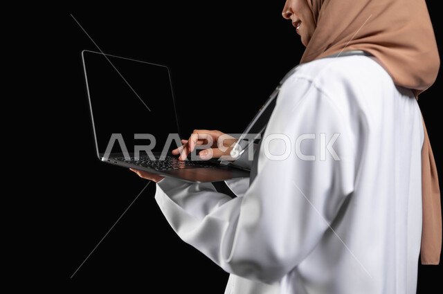 مفهوم استخدام أجهزة تقنيه حديثه، العمل في القطاع الصحي، بورتريه مقرب لطبيبة عربية خليجية سعودية مبتسمه محجبة  ترتدي المعطف الطبي تضغط بأصبعها على زر الحاسوب المحمول, خلفيه سوداء، محتوى حساس، استخدام حساس