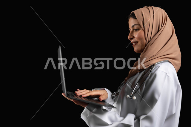 العمل في القطاع الصحي، مفهوم استخدام أجهزة تقنيه حديثه، بورتريه مقرب لطبيبة عربية خليجية سعودية مبتسمه محجبة  ترتدي المعطف الطبي تحمل بيدها الحاسوب المحمول, خلفيه سوداء، محتوى حساس، استخدام حساس
