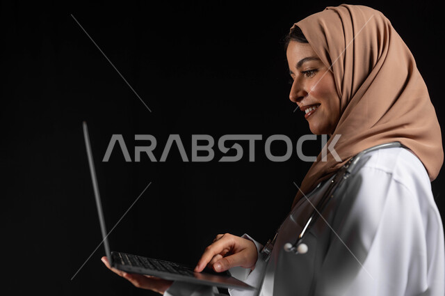 العمل في القطاع الصحي، مفهوم استخدام أجهزة تقنيه حديثه، بورتريه مقرب لطبيبة عربية خليجية سعودية مبتسمه محجبة  ترتدي المعطف الطبي تضغط بأصبع الابهام على زر الحاسوب المحمول, خلفيه سوداء، محتوى حساس، استخدام حساس