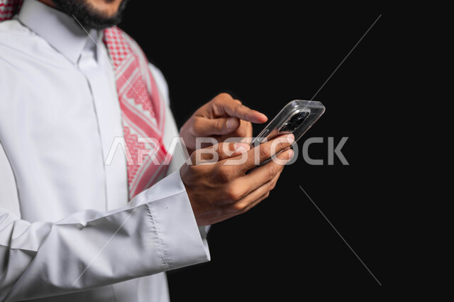 مفهوم الاناقة والاهتمام بالمظهر الخارجي، استخدام الهاتف المحمول، بورتريه لشاب عربي خليجي سعودي مبتسم يرتدي الزي التقليدي والشماغ  يحمل هاتف محمول، خلفية سوداء، محتوى حساس، استخدام حساس