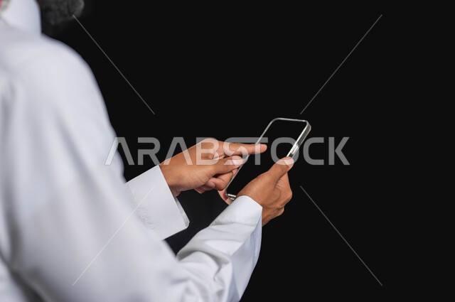 استخدام أجهزة تقنية حديثة، بورتريه مقرب لشاب عربي خليجي سعودي يرتدي الزي التقليدي يستخدم الهاتف المحمول، خلفية سوداء، محتوى حساس، استخدام حساس