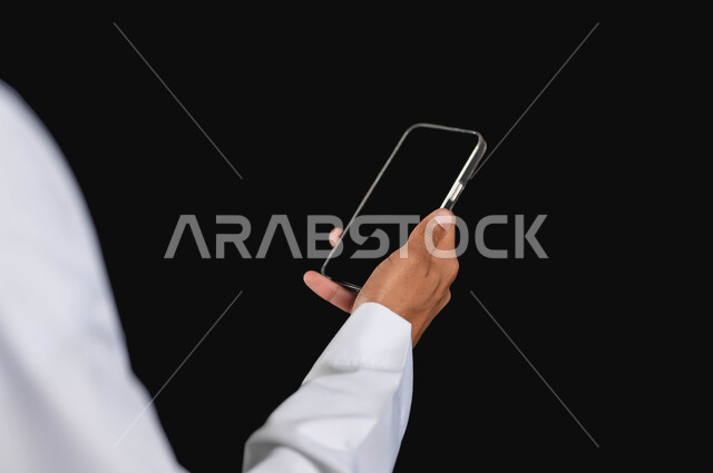 استخدام الأجهزة التقنية الحديثه، العمل عن بعد عبر الهاتف المحمول، بورتريه مقرب لرجل عربي خليجي سعودي يرتدي الزي التقليدي يحمل الهاتف المحمول،  خلفية سوداء، محتوى حساس، استخدام حساس