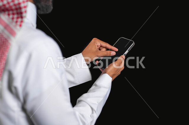 Using modern technical devices and browsing the Internet, a close-up portrait of a Saudi Gulf Arab man wearing traditional clothing, using a mobile phone, black background, sensitive content, sensitive use