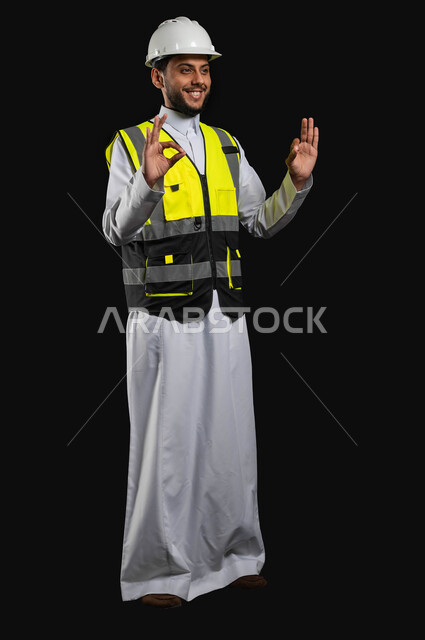 المهارة واتقان العمل، الشعور بالقبول والموافقة والرضا، بورتريه لمهندس عربي خليجي سعودي يرتدي الزي التقليدي مع سترة الحمايه والخوذة يرفع اصبع الابهام للاعلى بايماءات علامة الجوده، صورة الجسم بالطول الكامل، خلفية سوداء, محتوى حساس، استخدام حساس