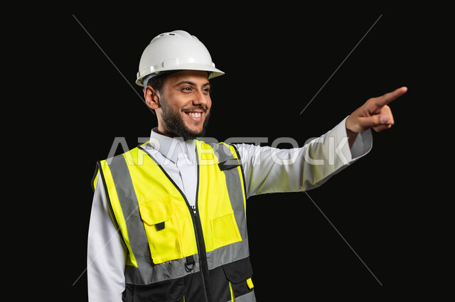 Looking with smiling gestures, pointing at something, working in the engineering sector, close-up portrait of a smiling Saudi Arabian Gulf engineer wearing a traditional uniform with a protective vest raising his hand towards a specific thing, black background, sensitive content, sensitive use