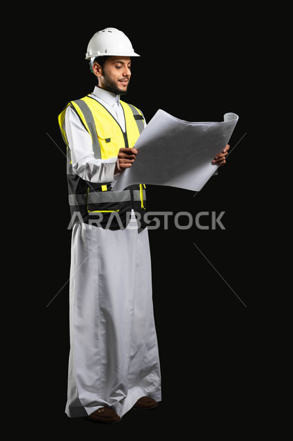 الدقة والالتزم بالعمل، مفهوم الانشاءات المعماريه، بورتريه مهندس عربي خليجي سعودي يرتدي الزي التقليدي مع سترة الحماية الخاصه يقوم بالعمل على مخططات المشروع، خلفية سوداء, محتوى حساس، استخدام حساس