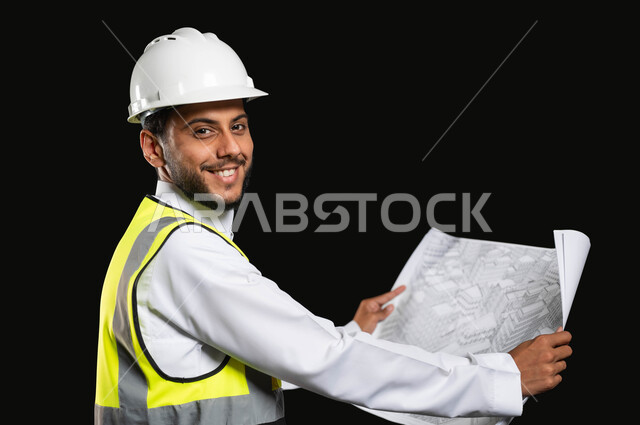 النظر الى الكاميرا بتعابير السرور، العمل في القطاع الهندسي، مفهوم ادارة المشاريع وتدقيقها، بورتريه مقرب لمهندس عربي خليجي سعودي يرتدي الزي التقليدي مع سترة الحماية والخوذة يقوم بمتابعة الاعمال ومخططات المشروع، خلفية سوداء, محتوى حساس، استخدام حساس