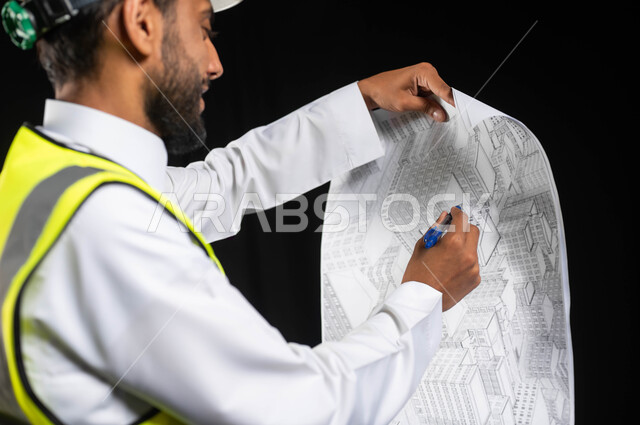 العمل في القطاع الهندسي،  مفهوم ادارة المشاريع وتدقيقها، بورتريه مقرب لمهندس عربي خليجي سعودي يرتدي الزي التقليدي مع سترة الحماية والخوذة يقوم برسم مخططات المشروع، خلفية سوداء,محتوى حساس، استخدام حساس