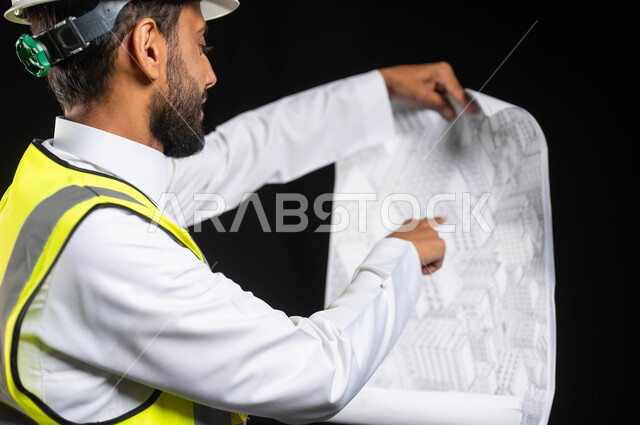 العمل في القطاع الهندسي، مفهوم ادارة المشاريع وتدقيقها، بورتريه مقرب لمهندس عربي خليجي سعودي يرتدي الزي التقليدي مع سترة الحماية والخوذة يقوم بمتابعة الاعمال ومخططات المشروع، خلفية سوداء,محتوى حساس، استخدام حساس