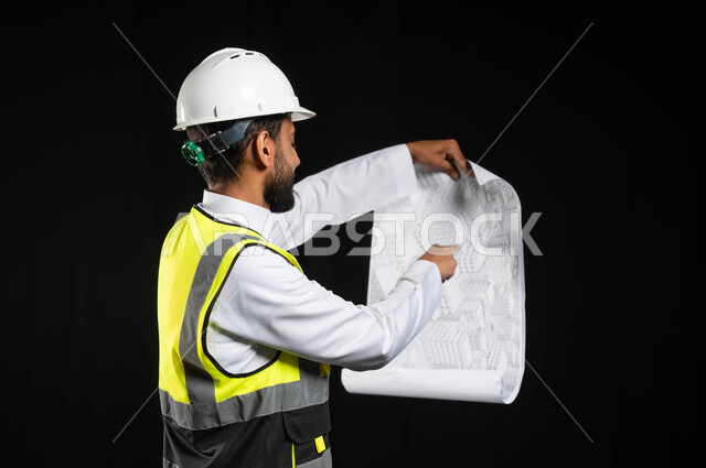 العمل في القطاع الهندسي،  مفهوم ادارة المشاريع وتدقيقها، بورتريه مقرب لمهندس عربي خليجي سعودي يرتدي الزي التقليدي مع سترة الحماية والخوذة يقوم بمتابعة الاعمال ومخططات المشروع، خلفية سوداء,محتوى حساس، استخدام حساس