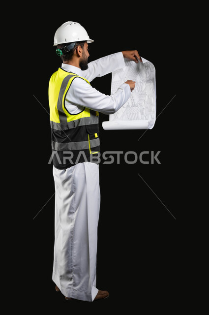 العمل في القطاع الهندسي،  مفهوم ادارة المشاريع وتدقيقها، بورتريه لمهندس عربي خليجي سعودي يرتدي الزي التقليدي مع سترة الحماية والخوذة يقوم بمتابعة الاعمال ومخططات المشروع، صورة الجسم بالطول الكامل، خلفية سوداء,محتوى حساس، استخدام حساس