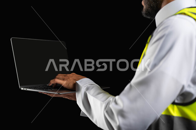 Working in the engineering sector, the concept of project management and auditing, a close-up portrait of a Saudi Gulf Arab engineer wearing traditional clothing working on a laptop, black background, sensitive content, sensitive use