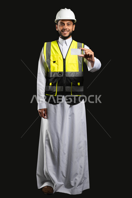 العمل في القطاع الهندسي، بورتريه لمهندس عربي خليجي سعودي يرتدي الزي التقليدي مع سترة الحماية والخوذة يحمل بيده كرت خاص ابيض، ادارة المشاريع وتدقيقها، خلفية سوداء, صورة الجسم بالطول الكامل، محتوى حساس، استخدام حساس