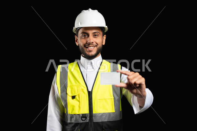 مهن ووظائف هندسية, العمل في القطاع الهندسي، بورتريه مقرب لمهندس عربي خليجي سعودي يرتدي الزي التقليدي مع سترة الحماية والخوذة يحمل بيده كرت خاص ابيض، ادارة المشاريع وتدقيقها، خلفية سوداء,محتوى حساس، استخدام حساس