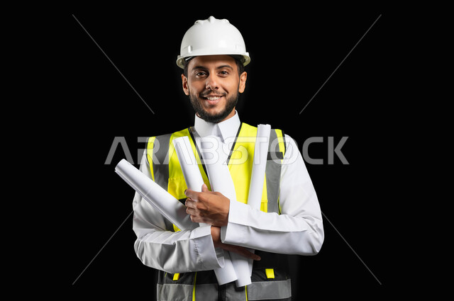النظر إلى الكاميرا بإيماءات السرور، الاهتمام بالتطور العمراني بسواعد المهندسين السعوديين، بورتريه مقرب لمهندس عربي خليجي سعودي يرتدي الزي التقليدي مع سترة الحماية والخوذة يمسك بيديه مخططات المشروع ، مهن ووظائف سعودية, خلفية سوداء, محتوى حساس، استخدام حساس
