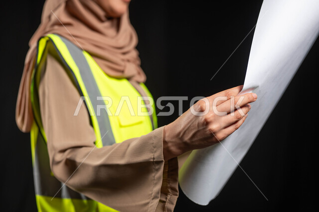 المخططات الهندسية المعمارية, بورتريه مهندسة معمارية عربية سعودية خليجية تحمل بيدها مخطط البناء ، مهندسة انشاءات محجبة ترتدي بدلة السلامة في العمل، لباس رسمي للعمل ، مشروع بناء ، خلفية سوداء,محتوى حساس, استخدام حساس