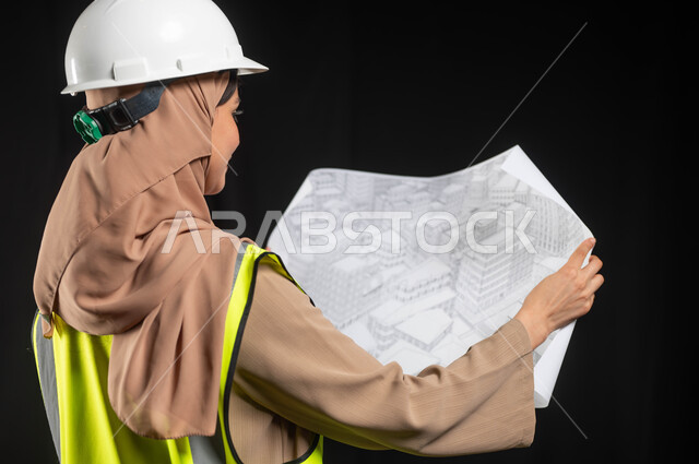 التصاميم المعمارية وتدقيق المخططات,العمل في المجال الهندسي, بورتريه من الخلف لمهندسة عربية خليجية سعودية محجبة ترتدي سترة الحماية والخوذة تمسك مخطط الإعمار, إدارة المشاريع الهندسية, مهن ووظائف نسائية, خلفية سوداء, محتوى حساس, استخدام حساس
