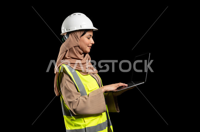 استخدام التكنولوجيا والتقنيات الحديثة، المتابعة والإشراف على العمل عبر جهاز اللابتوب المحمول, مهندسة عربية خليجية سعودية ترتدي سترة الحماية  والخوذة تستخدم الحاسوب، مشاركة النساء السعوديات في مجال الهندسة،بورتريه بخلفية سوداء, محتوى حساس, استخدام حساس