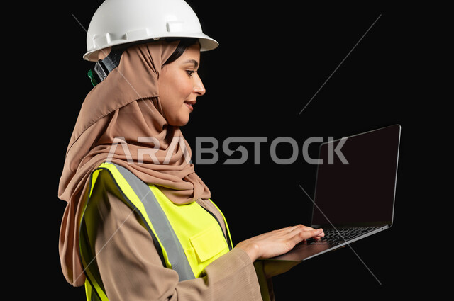 Managing engineering projects via the computer, studying the basics of the project using modern applications, integrating work with technology, a portrait from the side of a veiled Saudi Arabian Gulf engineer wearing a helmet and protective vest, holding a computer in her hand, black background, sensitive content, sensitive use