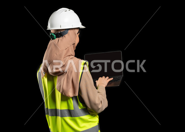 دمج العمل مع التكنولوجيا, دراسة أساسيات المشروع عبر التطبيقات والتقنيات الحديثة, إدارة المشاريع الهندسية, بورتريه  مقرب من الخلف لمهندسة عربية خليجية سعودية محجبة ترتدي الخوذة وسترة الحماية تمسك بيدها جهاز الحاسوب, خلفية سوداء، محتوى حساس، استخدام حساس