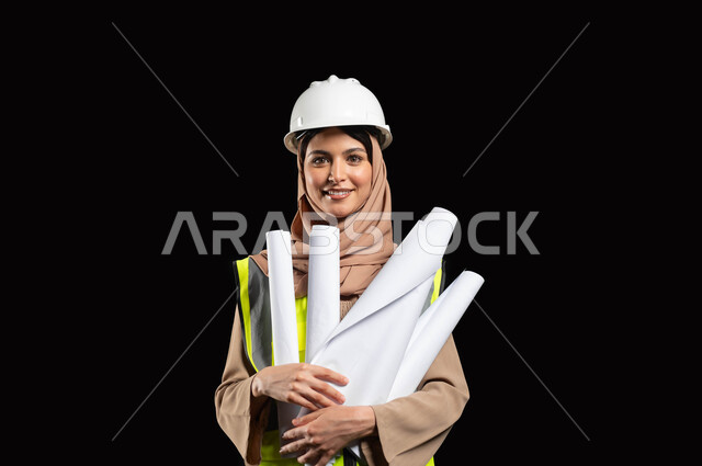 المشاريع الهندسية ومخططات الإعمار, النظر إلى الكاميرا بإيماءات السرور والفخر, بورتريه مقرب لفتاة عربية خليجية سعودية مبتسمة ترتدي سترة الحماية والخوذة تمسك مخطط الانشاء, العمل في القطاع الهندسي, خلفية سوداء, محتوى حساس, استخدام حساس