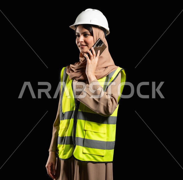مهن ووظائف نسائية هندسية سعودية, بورتريه مقرب مهندسة عربية خليجية سعودية محجبة ترتدي الخوذة وسترة الحماية تحمل بيدها الهاتف المحمول تجري مكالمة هاتفية، محتوى حساس, استخدام حساس