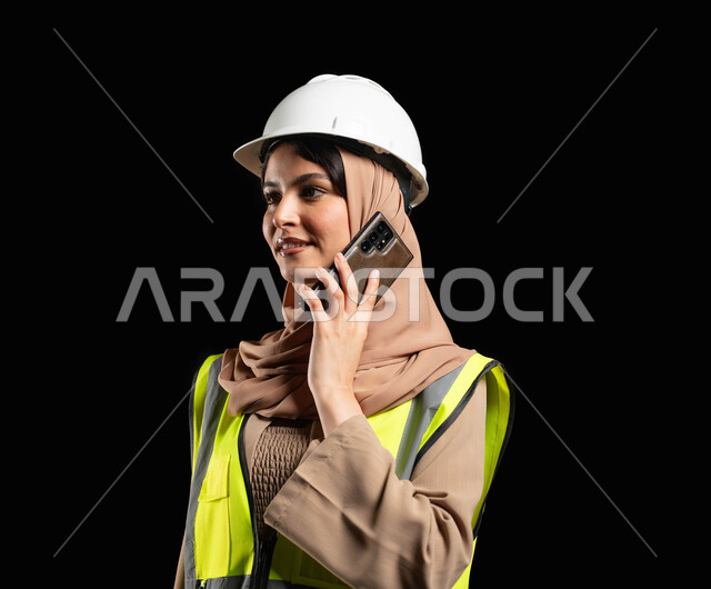 بناء و تطور المملكة العربية السعودية بأعمال أبناء الوطن, بورتريه مقرب لمهندسة عربية خليجية سعودية تجري مكالمة عبر الهاتف ترتدي سترة الحماية و الخوذة الخاصة بالاعمال الهندسية, خلفية سوداء, محتوى حساس, استخدام حساس