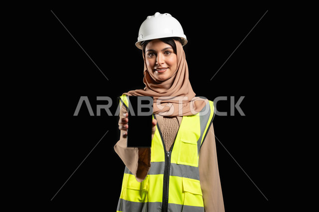 مهندسة عربية خليجية سعودية محجبة ترتدي خوذة الحماية والسترة, تحمل موبايل بشاشة سوداء ، إماءات الفرح و النجاح , الإشراف على المشاريع الهندسية في موقع العمل, خلفية سوداء، استخدام حساس, محنوى حساس