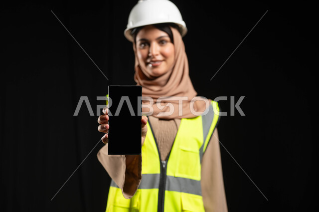 Saudi engineering jobs, close-up portrait of a Saudi Gulf Arab engineer wearing a protective helmet and jacket holding the phone with a black screen, black background, sensitive use, sensitive content