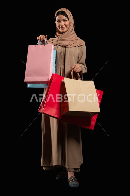 التسوق  في المراكز التجاريه السعودية, بورتريه لإمرأة عربيه خليجية سعوديه محجبة, ترتدي عباءه عصرية و حجاب و تحمل بيدها حقائب التسوق الملونة, صورة الجسم بالطول الكامل, خلفية سوداء محتوى حساس, استخدام حساس