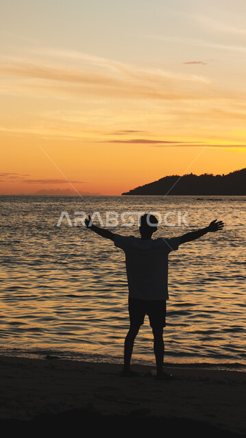 غروب الشمس فوق ساحل البحر، الواجهة البحرية والمناطق الطبيعية في المملكة العربية السعودية، النزهات والرحلات في فصل الصيف، قضاء وقت ممتع بتأمل مناظر الطبيعة الهادئة، صورة ظلية من الخلف لشاب عربي خليجي سعودي يقف على الشاطئ الرملي، سالويت