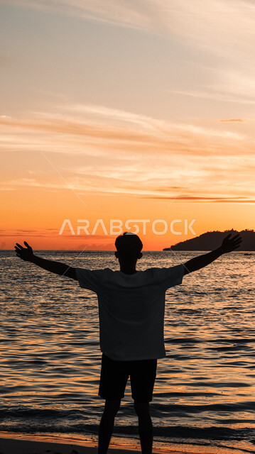 الواجهة البحرية والمناطق الطبيعية في المملكة العربية السعودية، النزهات والرحلات في فصل الصيف، قضاء وقت ممتع بتأمل مناظر الطبيعة الهادئة، منظر غروب الشمس فوق ساحل البحر، صورة ظلية من الخلف لشاب عربي خليجي سعودي يقف على الشاطئ الرملي، سالويت