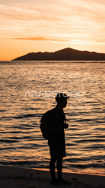 Picnics and trips in the summer, natural areas and the waterfront in the Kingdom of Saudi Arabia, spending a pleasant time contemplating tranquil nature views, a view of the sunset over the sea coast, a silhouette from behind of a young Saudi Gulf Arab man standing on the sandy beach, Salwit