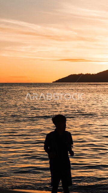 الرحلات والنزهات في فصل الصيف، المناطق الطبيعية والواجهة البحرية في المملكة العربية السعودية، قضاء وقت ممتع بتأمل مناظر الطبيعة الهادئة، منظر غروب الشمس فوق ساحل البحر، صورة ظلية من الخلف لشاب عربي خليجي سعودي يقف على الشاطئ الرملي، سالويت