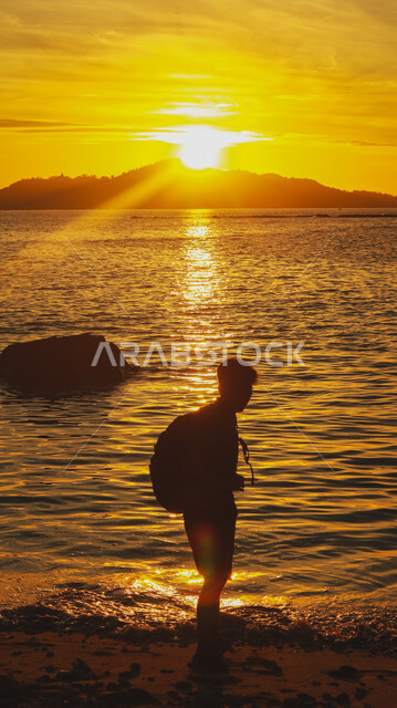Natural places and the waterfront in the Kingdom of Saudi Arabia, spending a pleasant time contemplating the tranquil landscapes, areas for excursions and picnics in the summer, a view of the sunset over the sea coast, a silhouette from behind of a young Saudi Gulf Arab man standing on the sandy beach, Silhouette