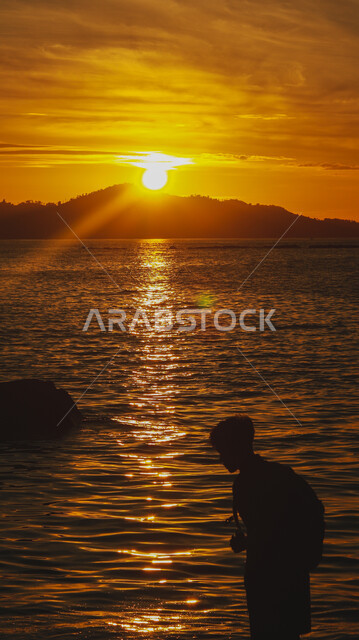 قضاء وقت ممتع بتأمل مناظر الطبيعة الهادئة، مناطق الرحلات والنزهات في الصيف، منظر غروب الشمس فوق ساحل البحر، صورة ظلية من الجانب لشاب عربي خليجي سعودي يقف على الشاطئ الرملي، الأماكن الطبيعية والواجهة البحرية في المملكة العربية السعودية، سالويت